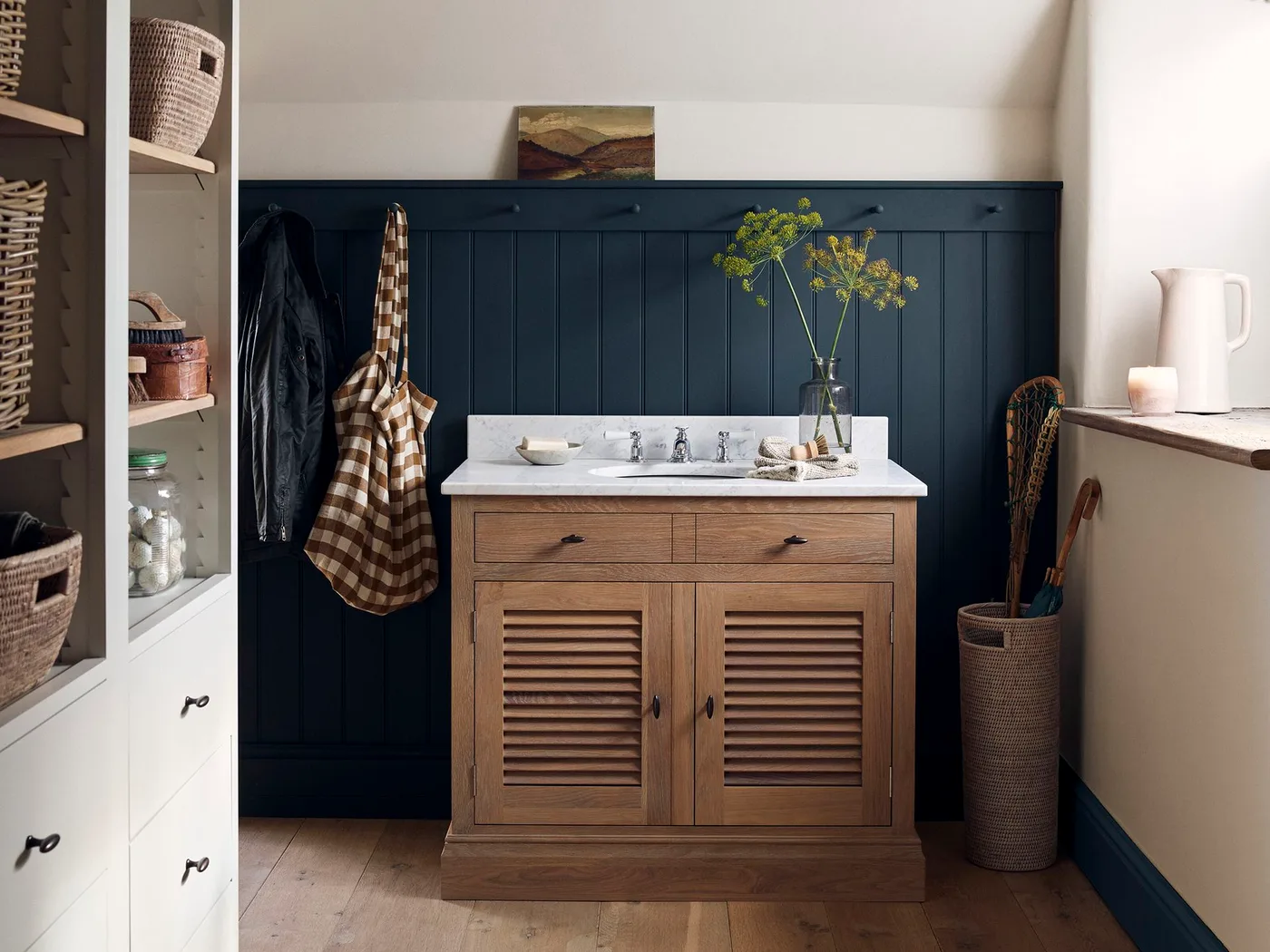 Small bathroom with navy vertical paneling, a wooden vanity, open shelves of baskets, wall hooks and a tall basket beside the window.