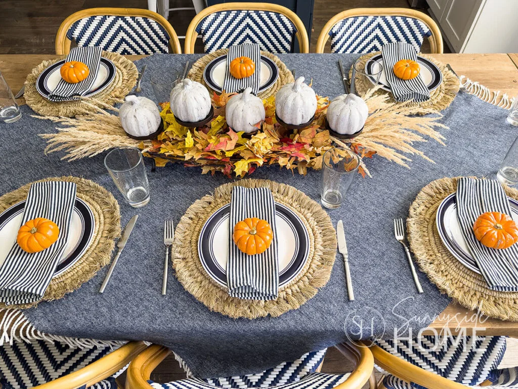 Small orange pumpkins top striped napkins around a blue-and-white dining table with woven chargers and pale pumpkins along the center