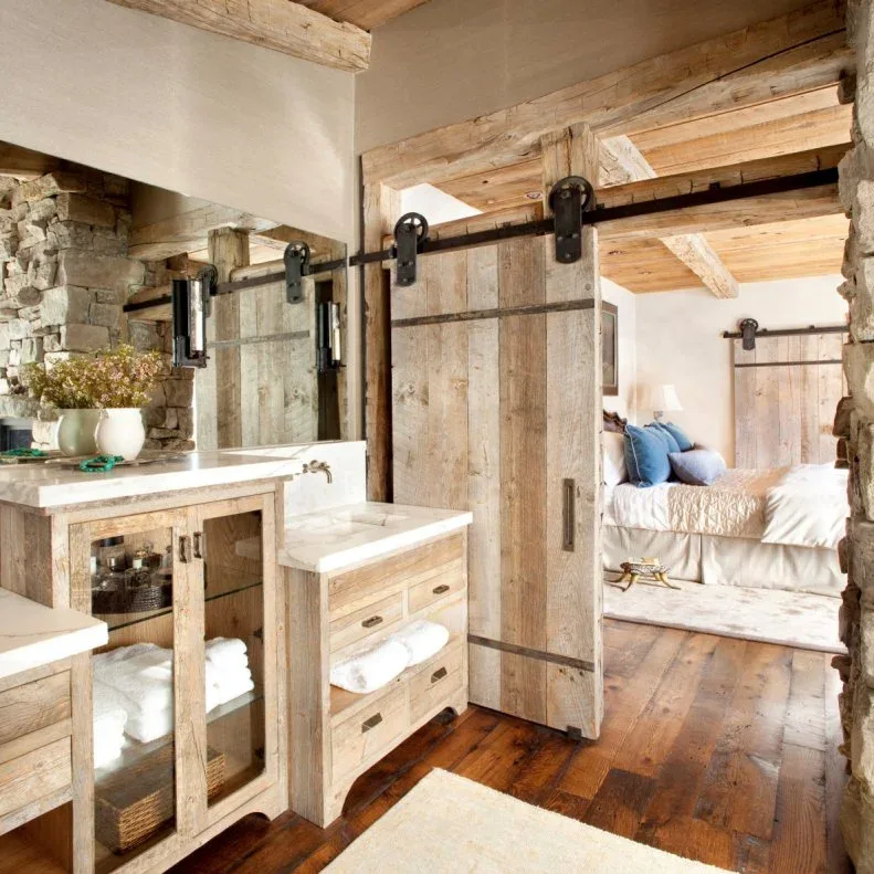 Rustic bathroom with wood vanities, rough masonry, ceiling beams and wood sliding door beside an adjoining bed