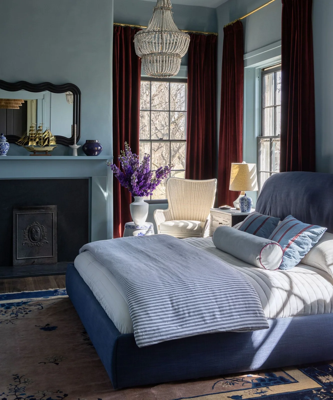 Dusty blue bedroom walls and fireplace surround beside a blue bed with pale bedding and red curtains.