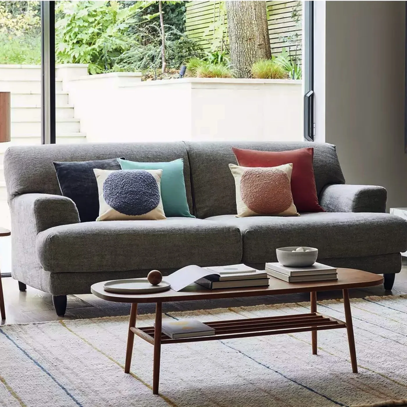 A gray sofa with blue, turquoise and red cushions sits near broad garden glazing behind a low wooden coffee table and pale rug