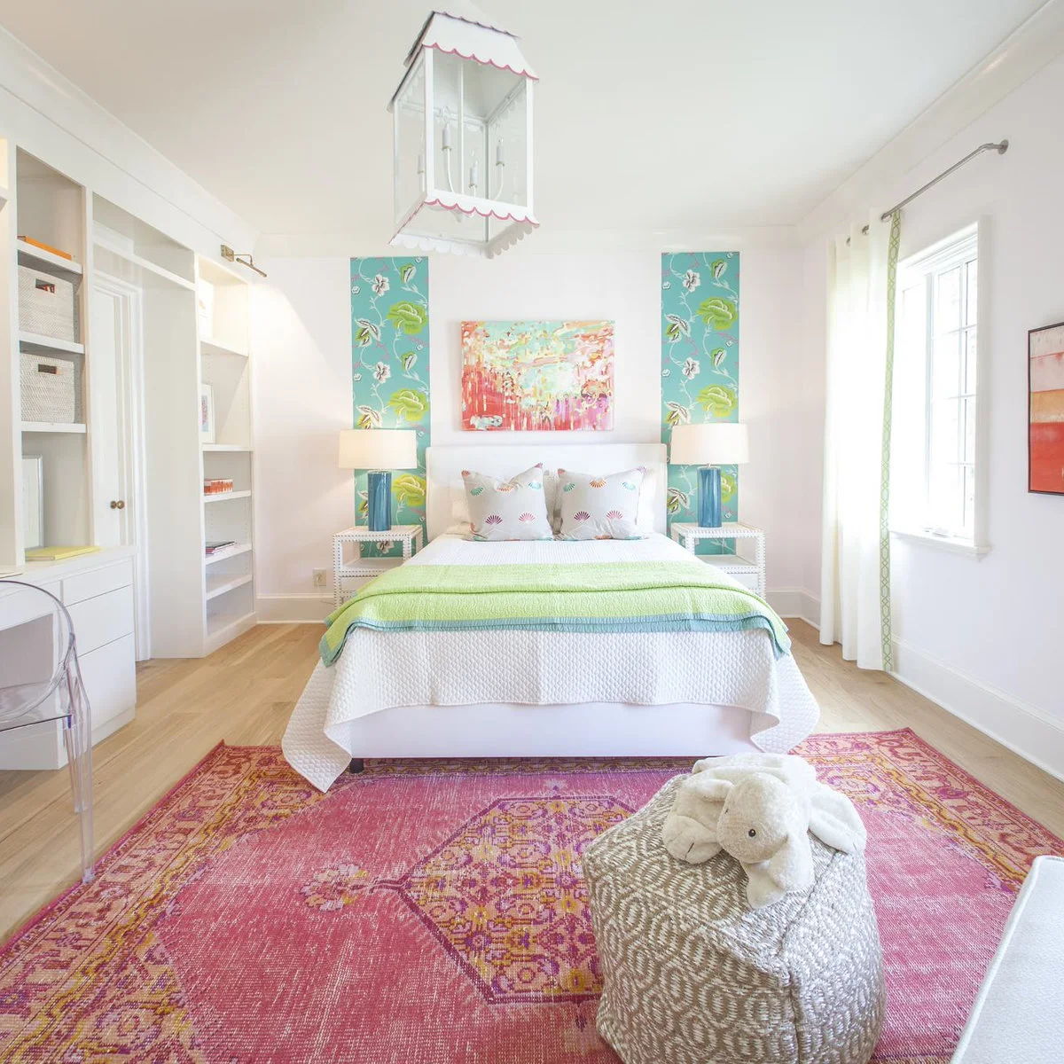 A pink and gold patterned rug extends in front of a white bed with a green throw, blue floral panels and a woven pouf.