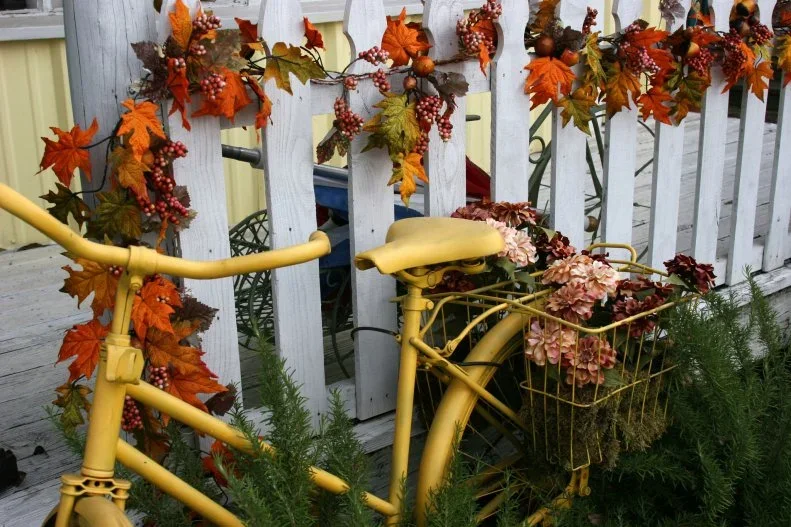 Orange leaves and berry-like clusters run along a pale porch rail behind a yellow bicycle and flower basket