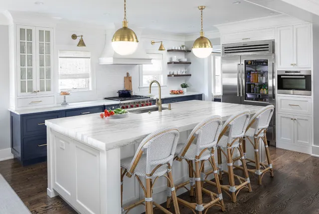 Blue lower cabinets surround a pale island with woven chairs and brass-toned pendants.
