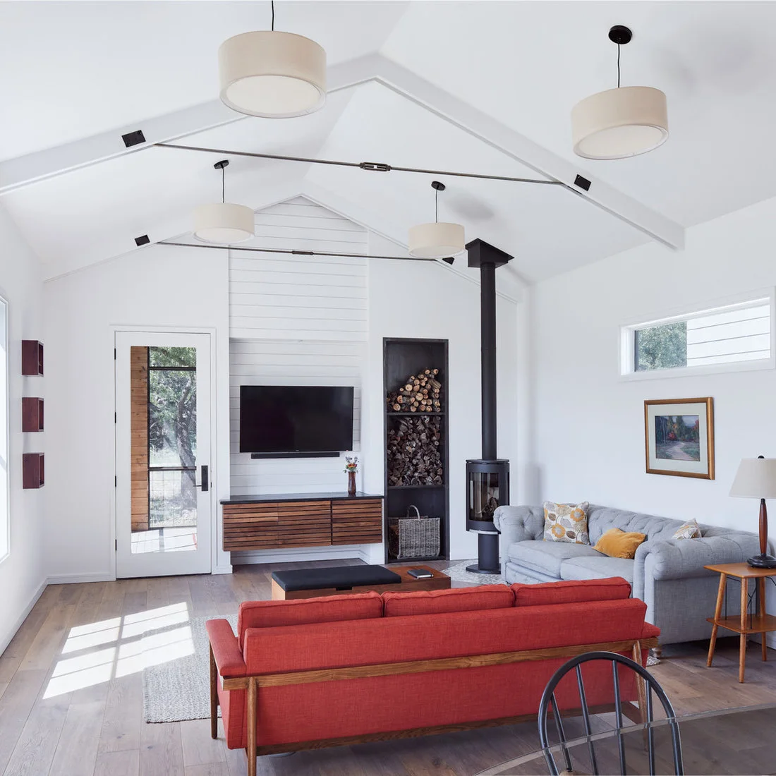 A white vaulted room has a slim black cylindrical stove, tall recessed log storage, gray and red sofas, a low wood cabinet and simple drum pendant shades.