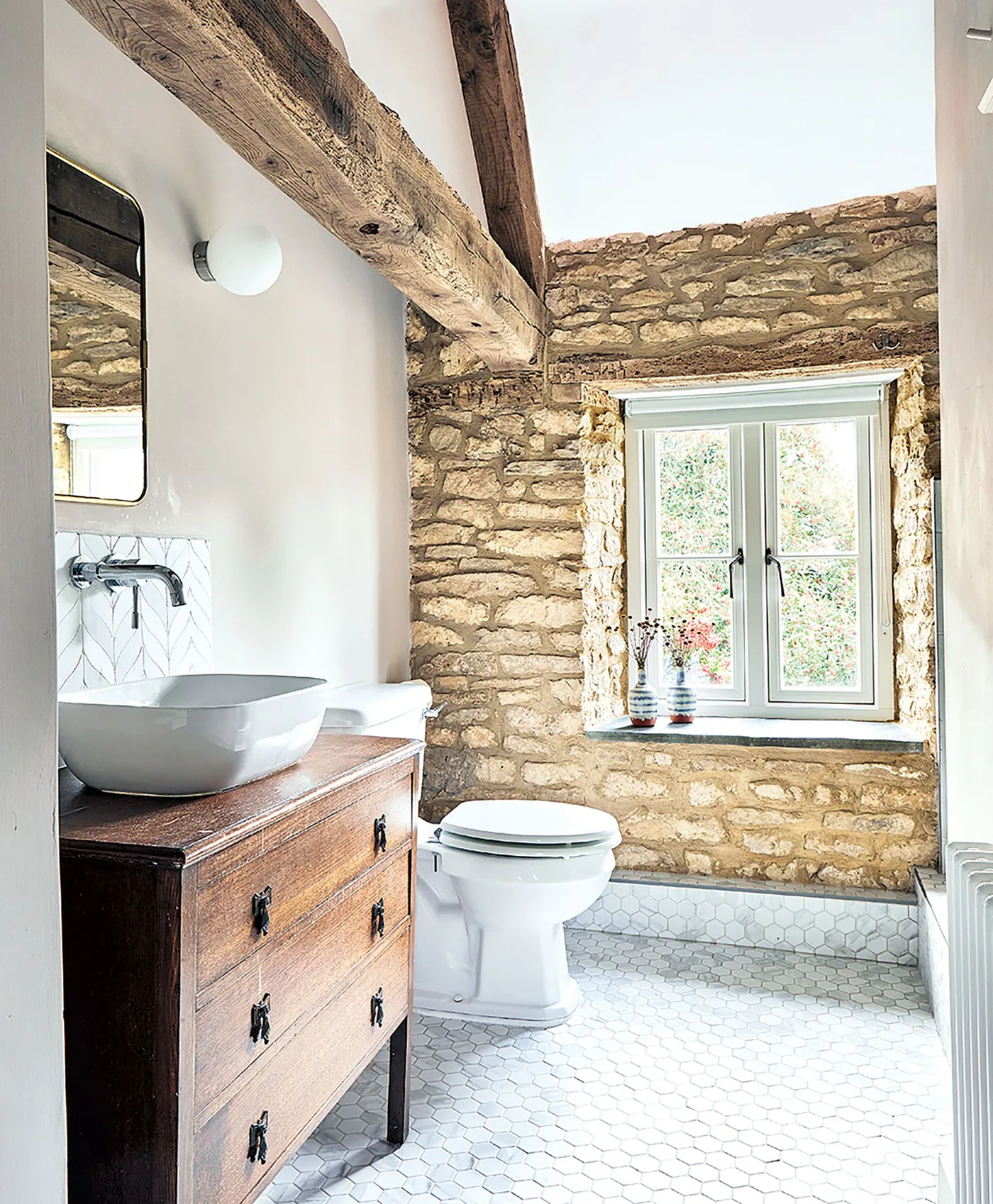 Bathroom with a rough stone wall, exposed wooden beams, wooden drawer washstand, white vessel basin and pale tiled floor.