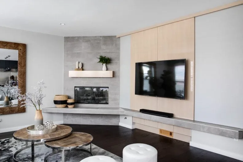 A gray living-room fireplace wall meets a pale wood television wall above low storage beside round nesting tables and a patterned rug