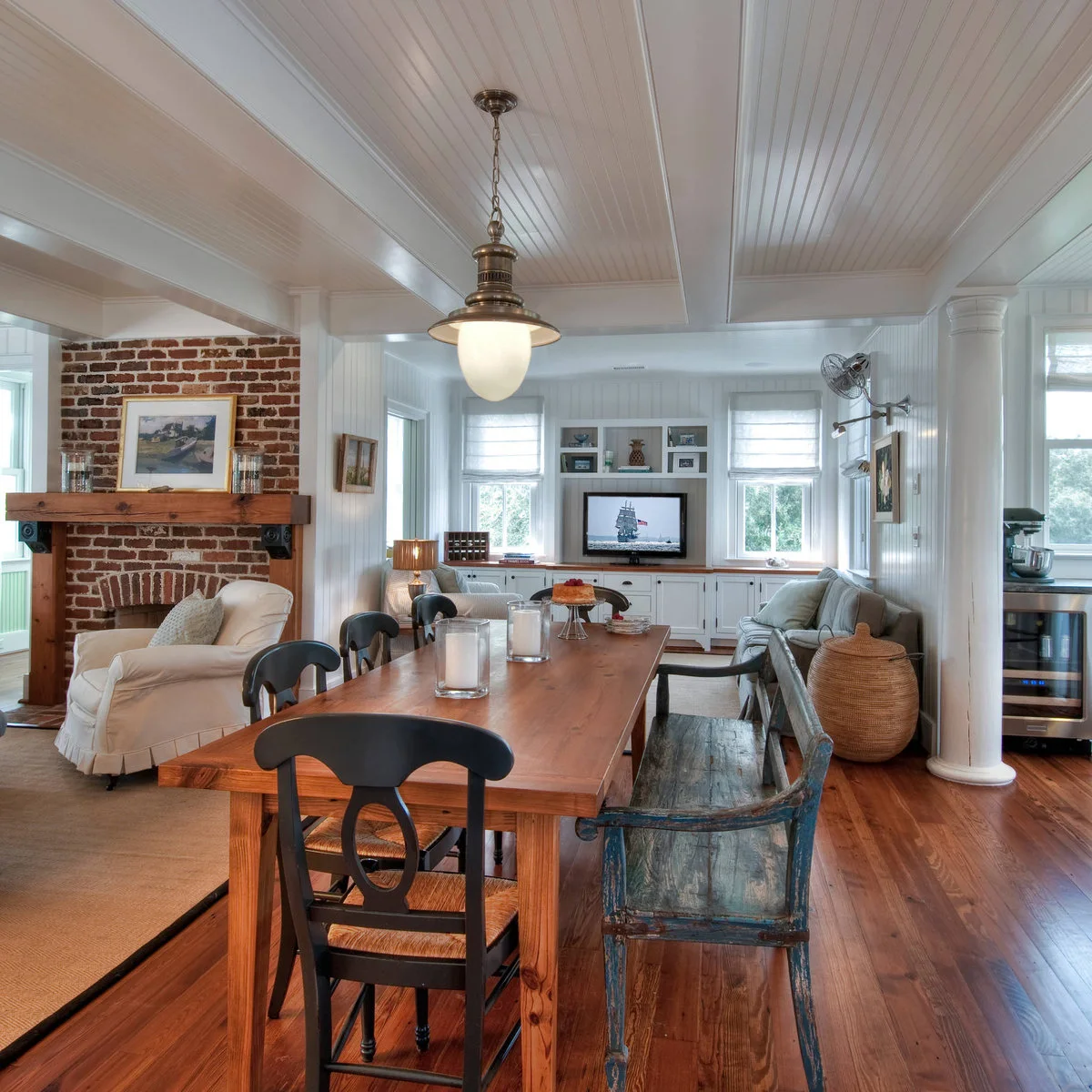 Long white paneled room with a wooden dining table, dark chairs, a gray sofa, brick fireplace and a white column beside the kitchen.