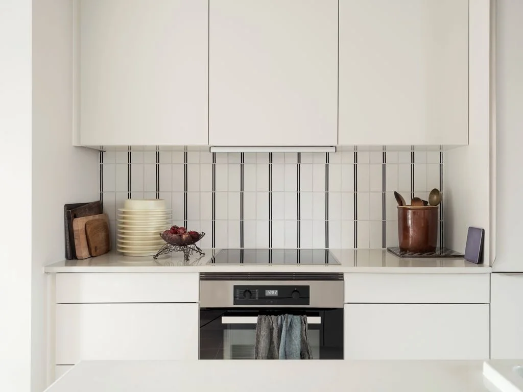 A compact white kitchen has flat upper and lower fronts, a small cooktop, an oven below and a pale backsplash crossed by fine dark stripes.