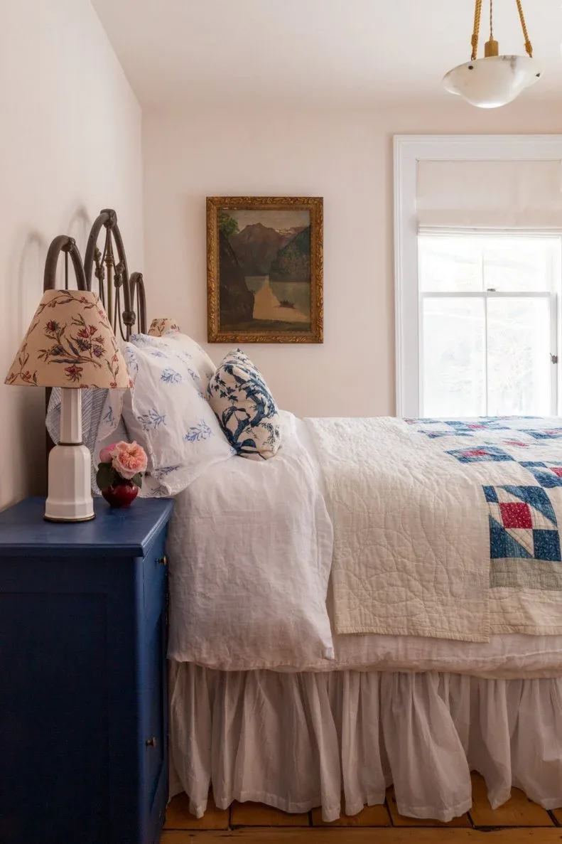 A dark metal bed has a colorful patchwork quilt, pale bedding and a blue bedside cabinet beneath a floral lamp shade.