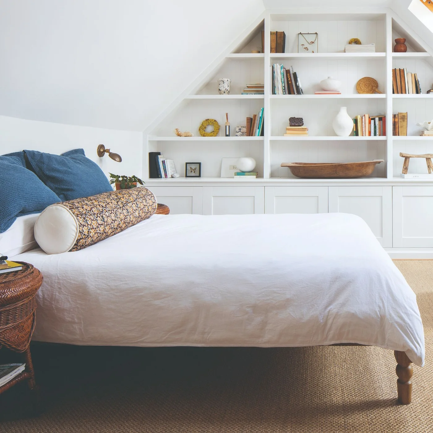 White shelves follow a triangular attic wall above closed cupboards beside a bed with blue pillows, a patterned bolster and a small woven bedside table.