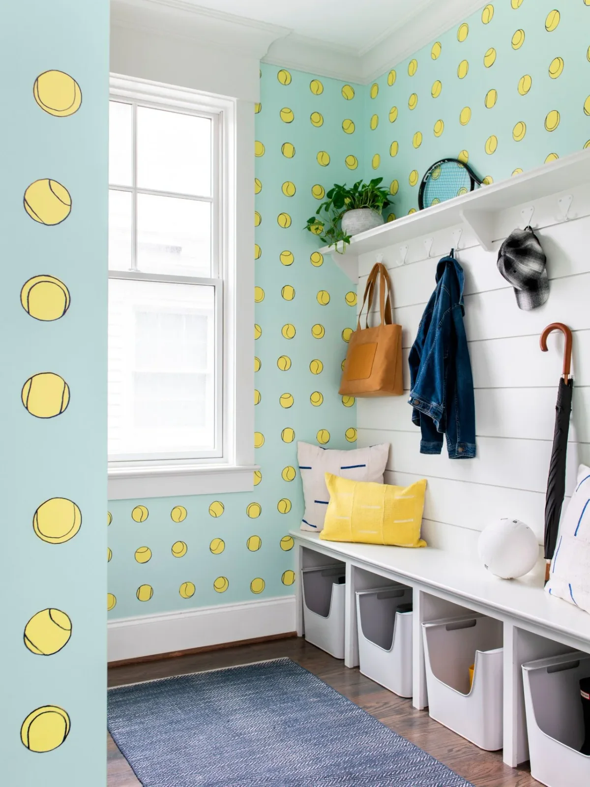 A white bench with gray storage bins and yellow pillows stands against tennis-ball wallpaper below a coat rail.
