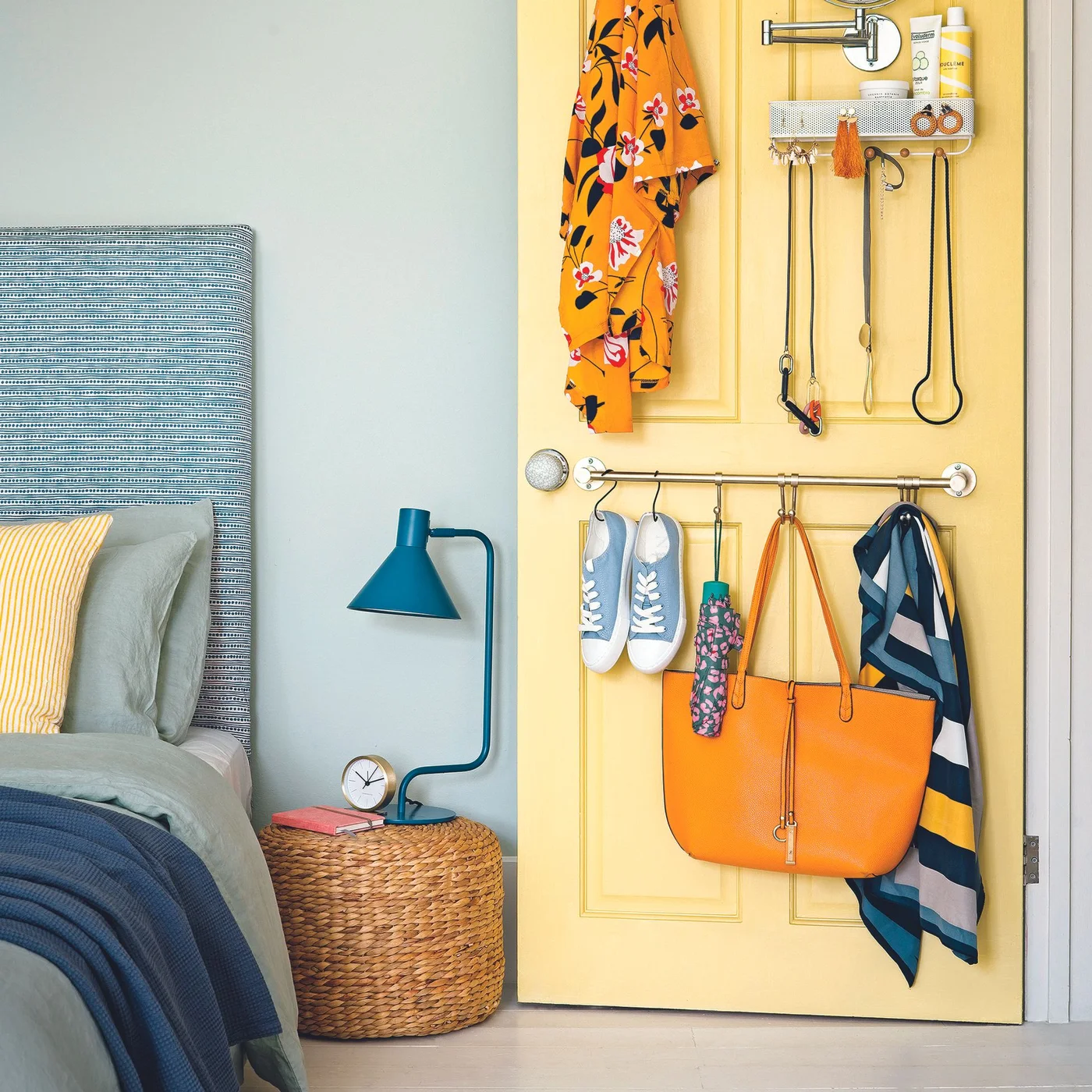A yellow bedroom door carries rails, hooks and a small organizer for shoes, bags, jewelry and toiletries beside a bed and a woven cylindrical table.