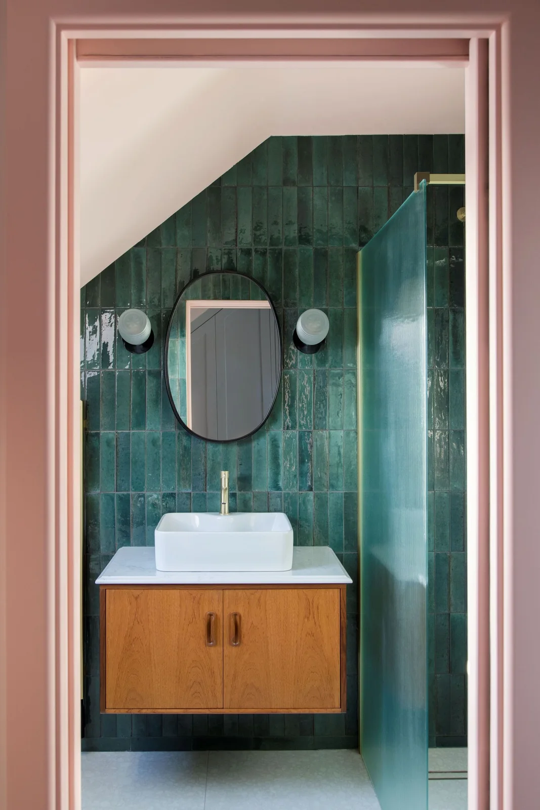 A round dark-framed bathroom mirror sits between two globe lights against deep green tiles above a floating wood vanity within a pink doorway