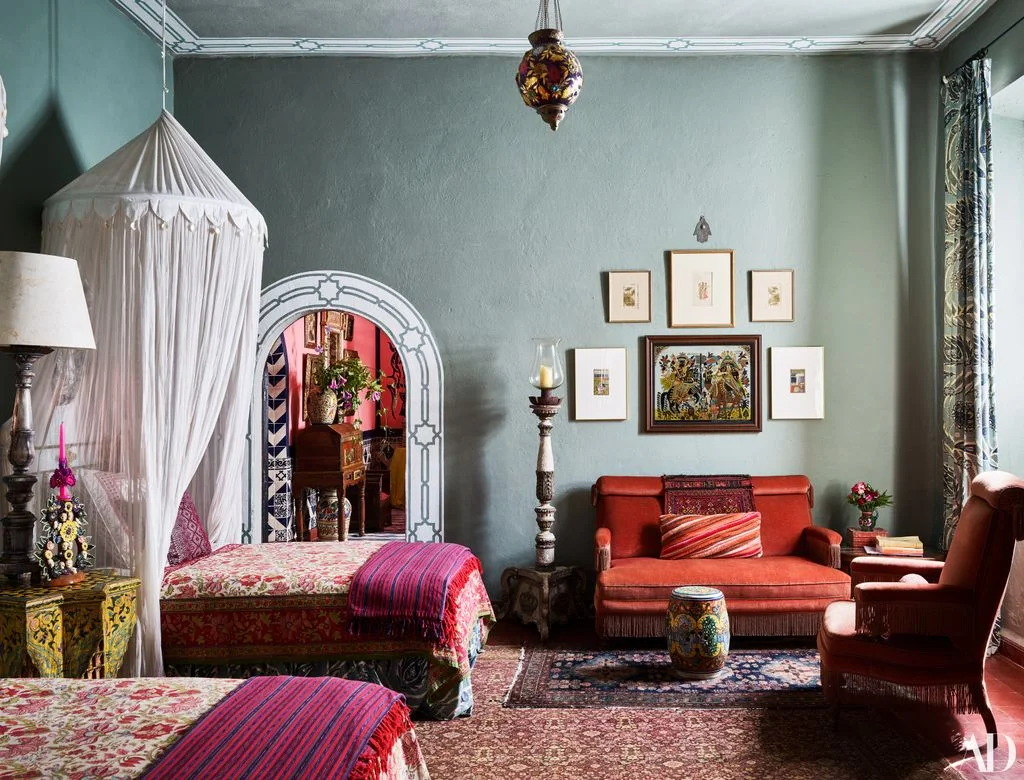 Two patterned canopy beds with bright striped throws beside an orange settee and a small framed picture group on a pale blue wall.