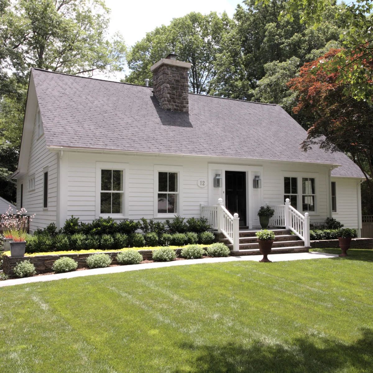 A white Cape Cod house with dark shrubs behind lighter rounded planting in a brick-edged front bed.