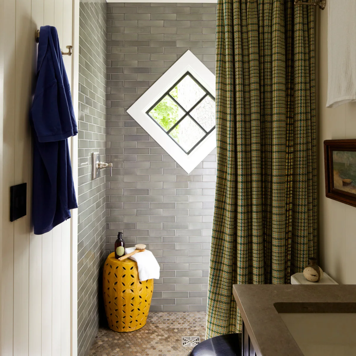 A green-and-gold checked shower curtain hangs beside gray tiles, a diamond-shaped window and a yellow stool.