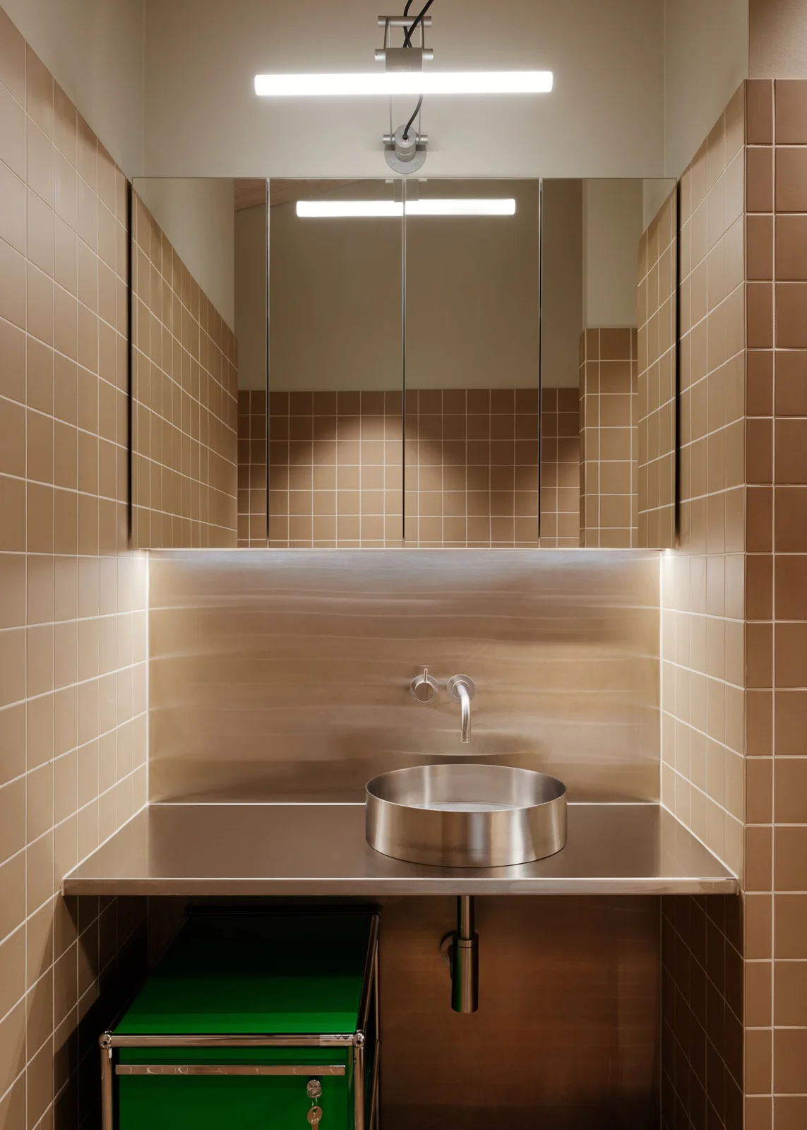 A metal counter and round basin sit beneath mirrored cupboards between warm beige tiled walls with a green storage unit below.
