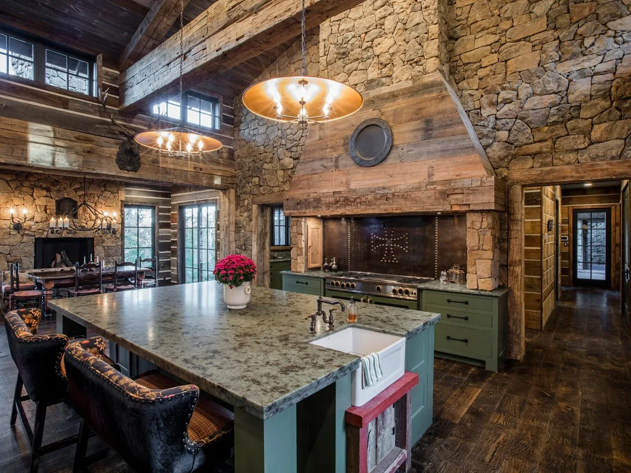 Green kitchen cabinets and a broad island sit beneath a timber hood among rough stone walls and exposed beams.