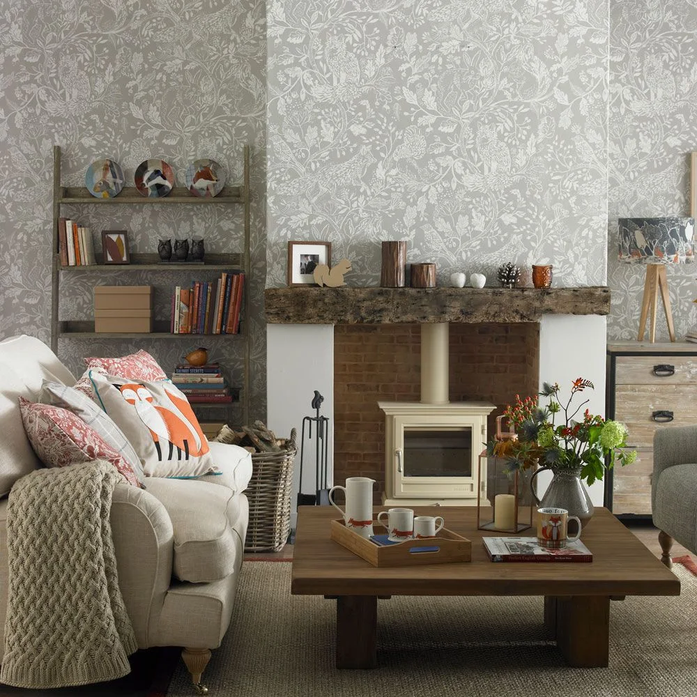 Pale woodland-patterned wallpaper surrounds a rough wood mantel above a cream stove beside pale seating and a square wood table.