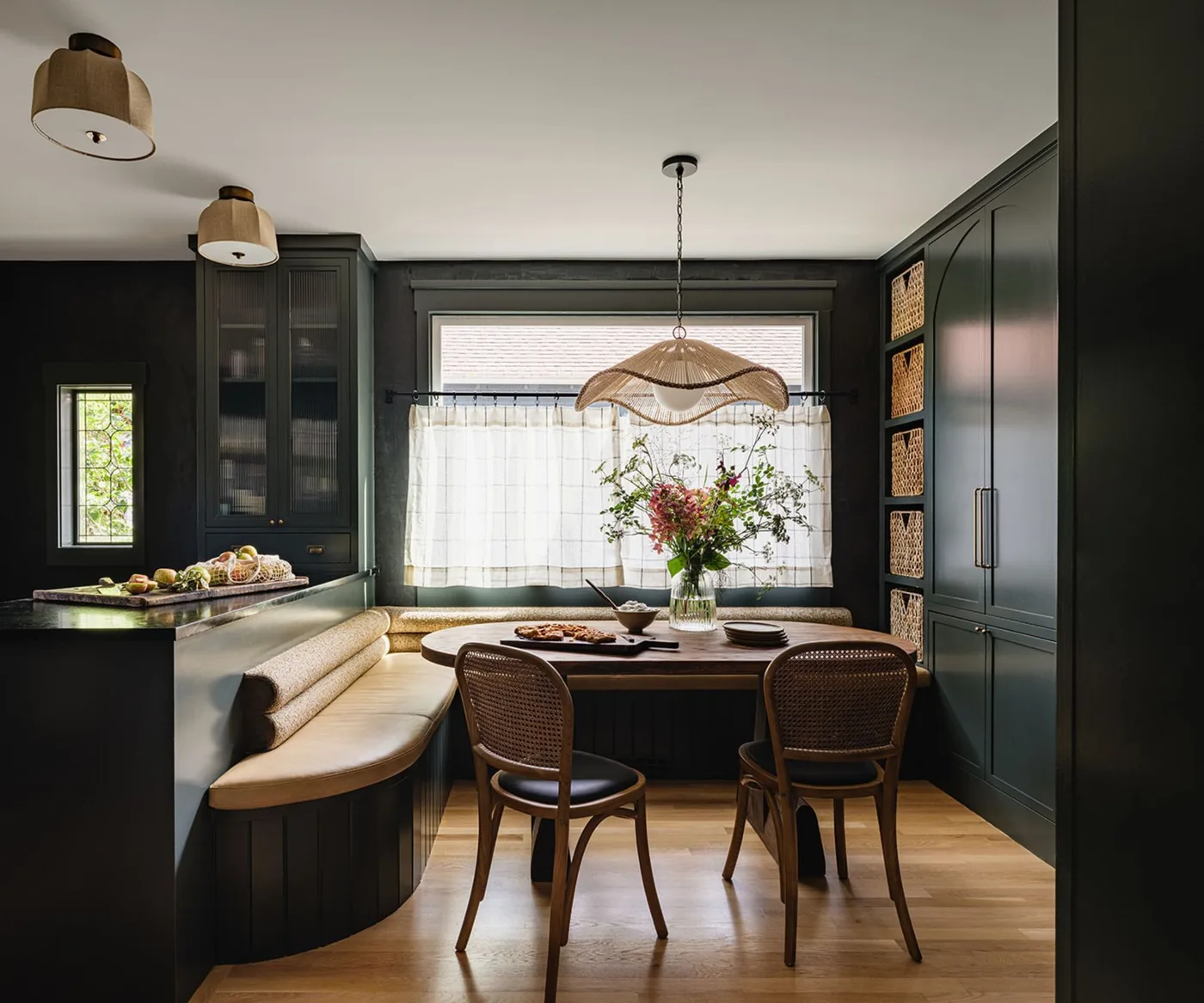 Dark green paneled dining nook with a pale curved banquette, round wooden table and two wooden chairs with woven backs.