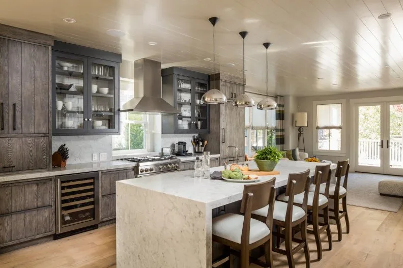 Dark wood lower and tall kitchen cabinets mix with black glass-front uppers, with a pale waterfall island and light wood flooring.