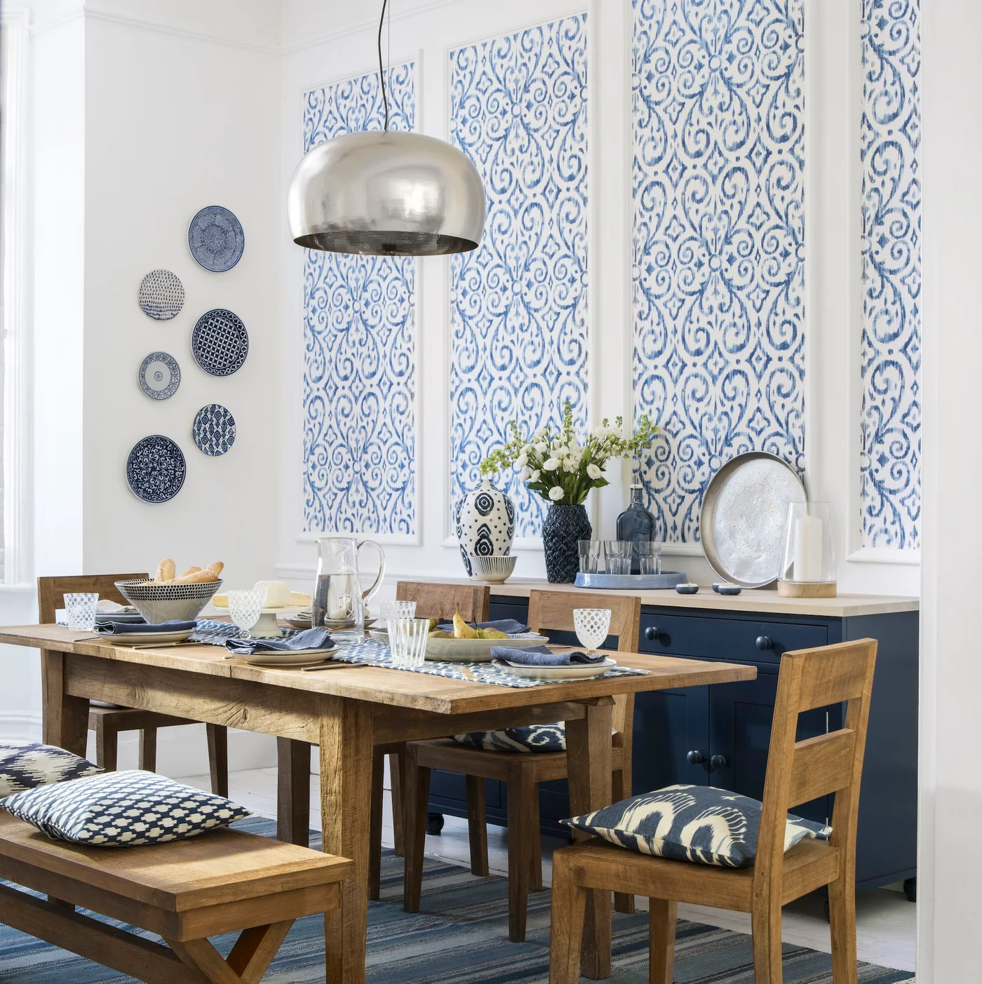 A navy sideboard with a pale top sits below blue-and-white wallpaper panels behind a timber dining table and chairs.