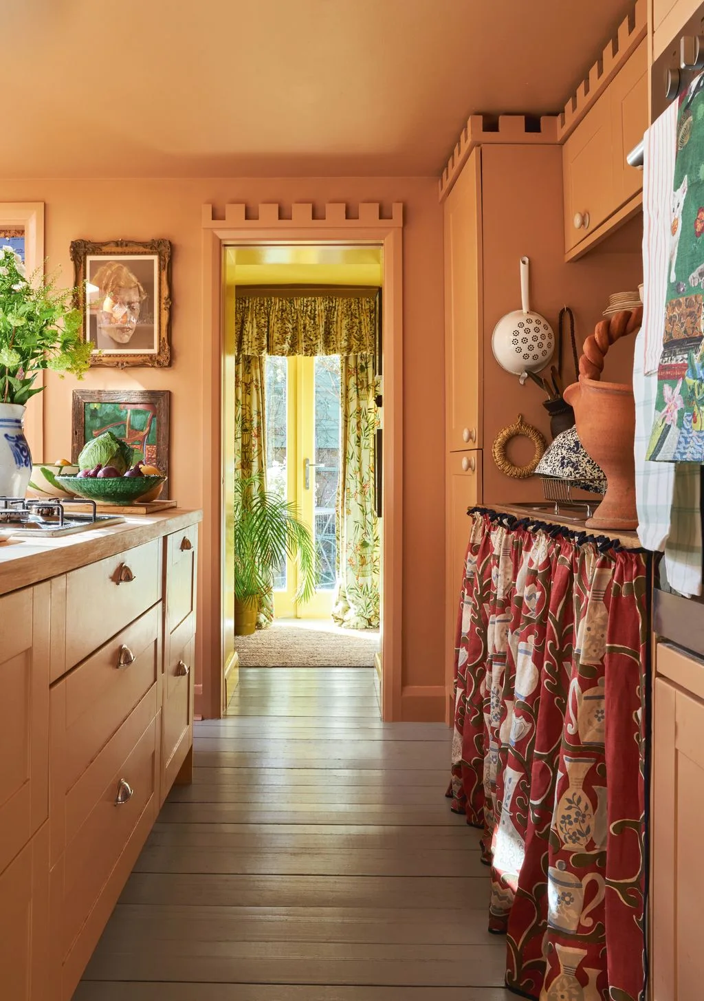 Peach walls and cabinets with crenellated upper edges surround framed pictures, a patterned cabinet skirt, hanging utensils and printed doorway curtains