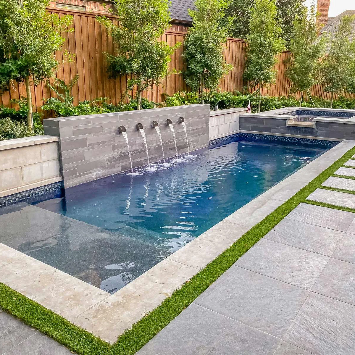 A narrow blue pool and raised spa sit beside a long gray wall with arched water spouts, planting and a paved lounging area.