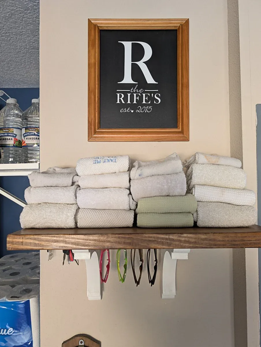 Wooden shelf with folded towels and sunglasses hanging from hooks beneath it on a garage wall.