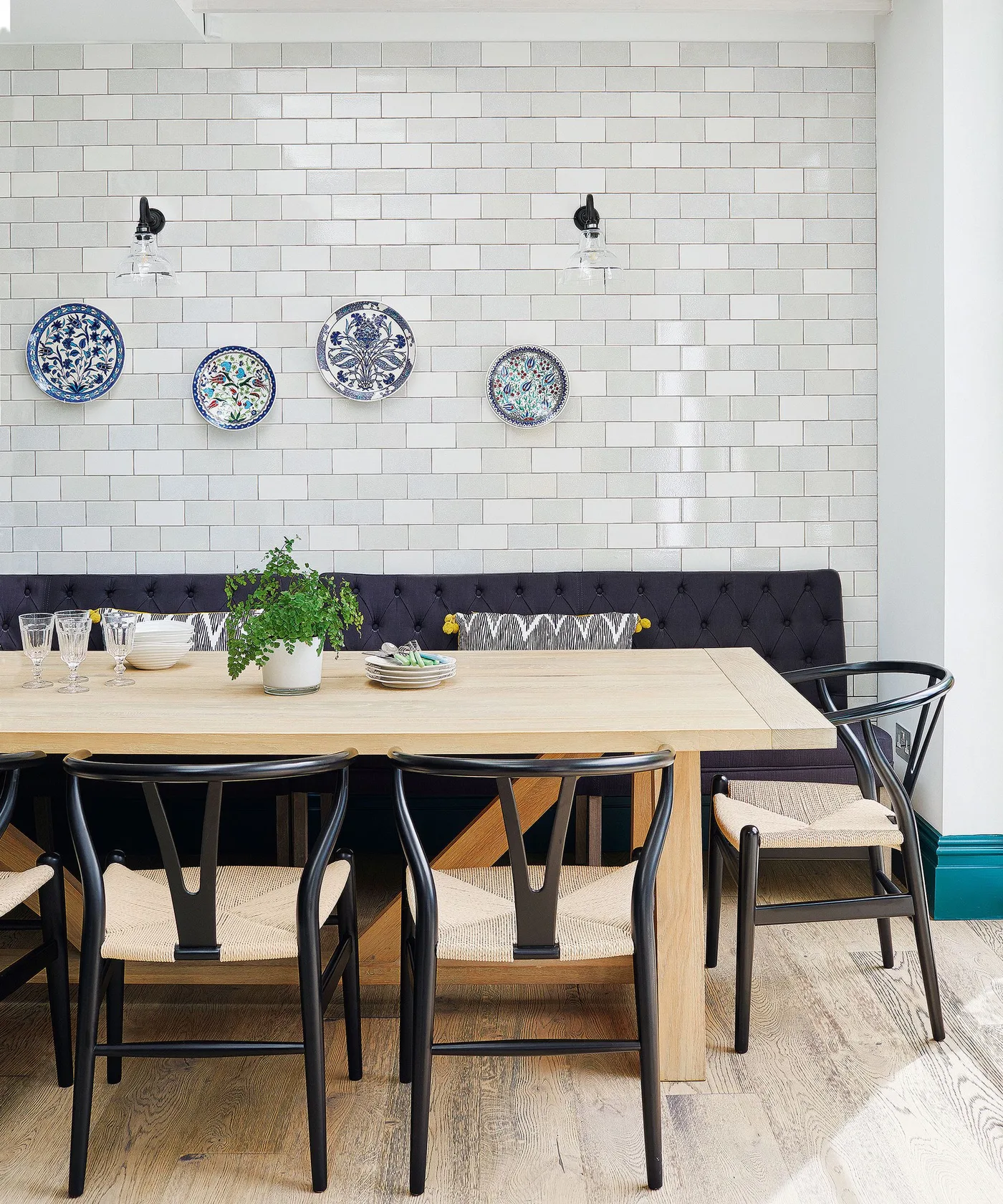 Four blue-patterned decorative plates on a white tiled wall above a dark blue buttoned bench, with a wooden table, black chairs, and two dark wall lights.