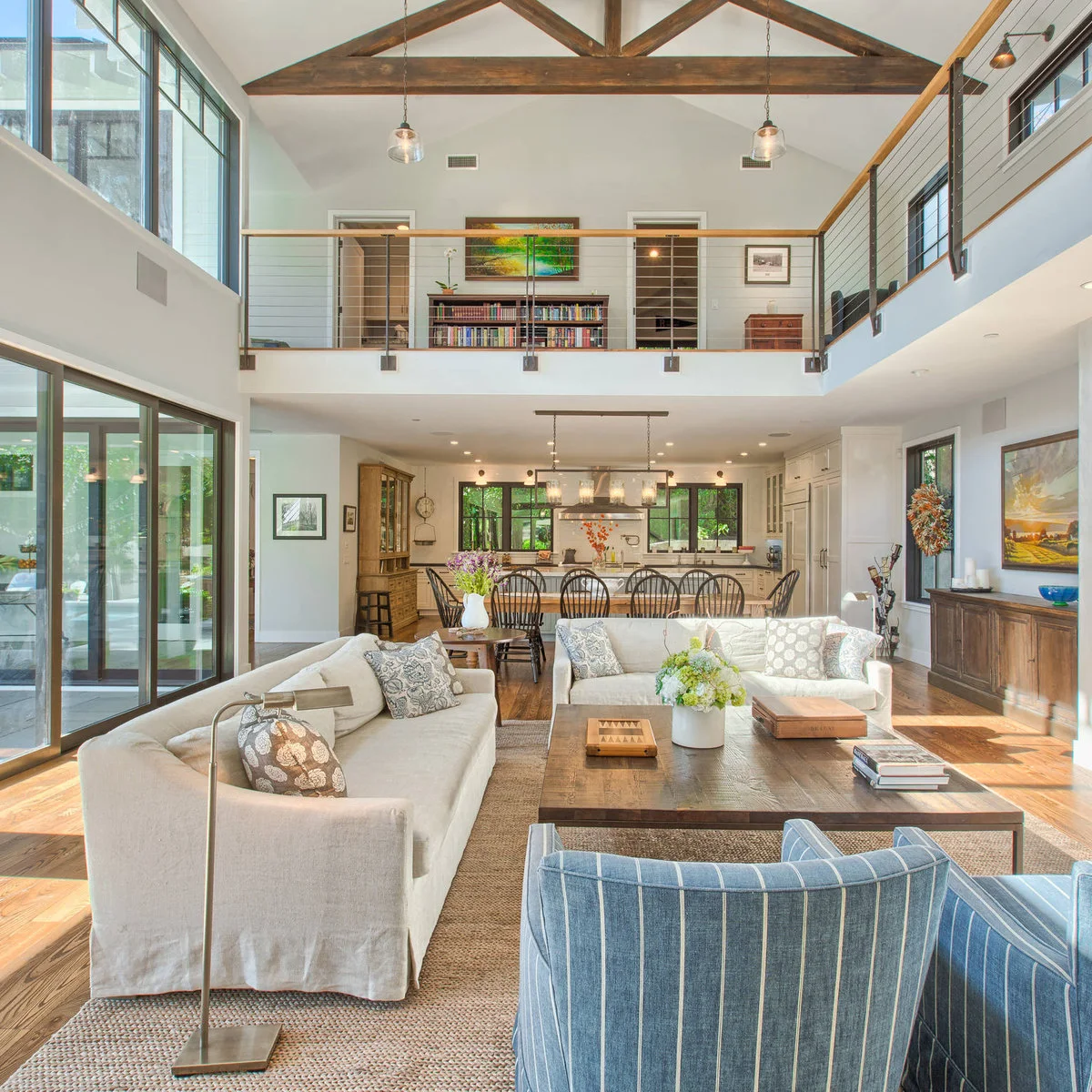 A tall room with exposed roof trusses, a mezzanine, cream sofas, blue striped chairs and a broad low wood table.