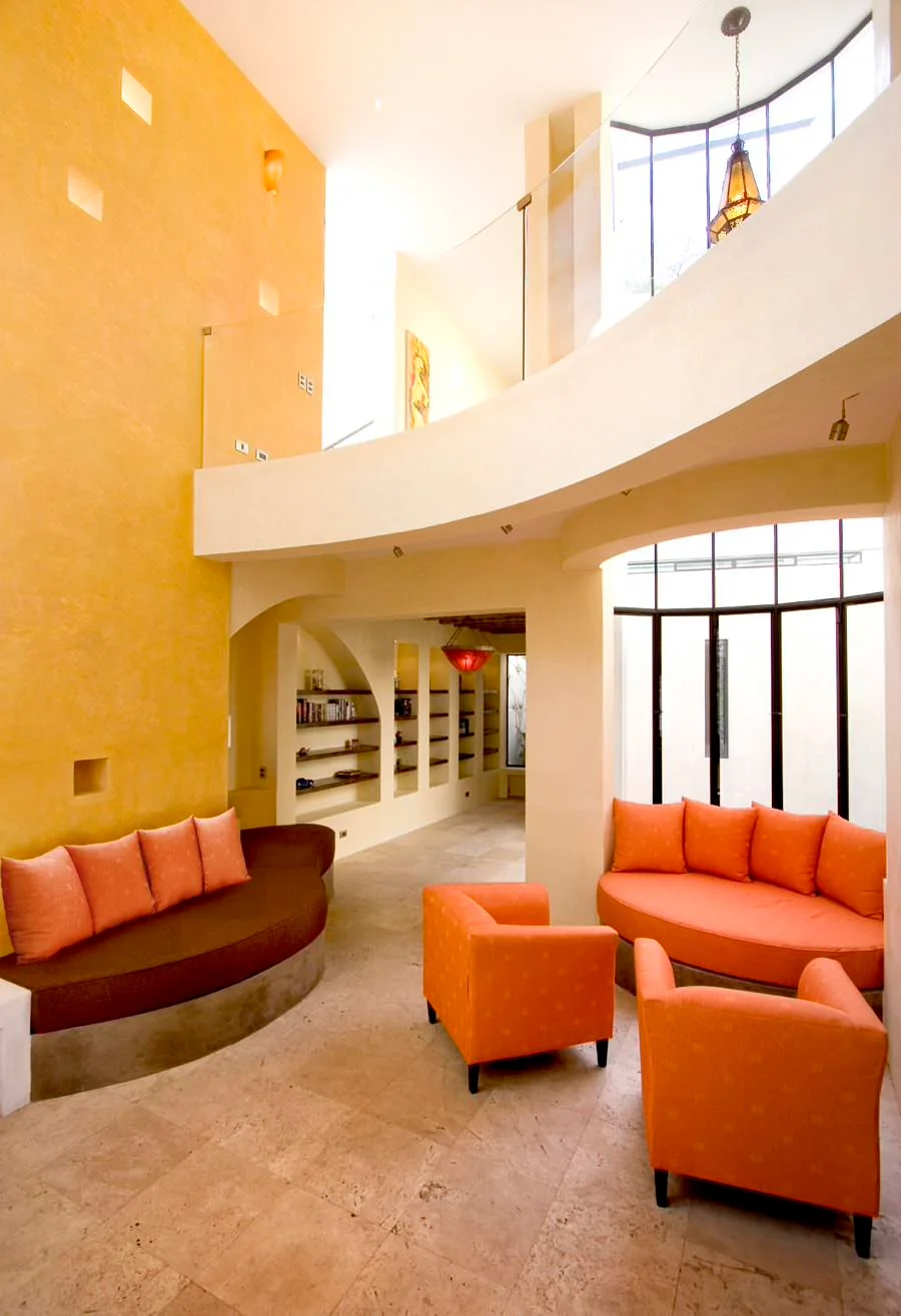 Curved built-in brown and orange sofas with orange armchairs in a tall yellow room beside large black-framed glazing.