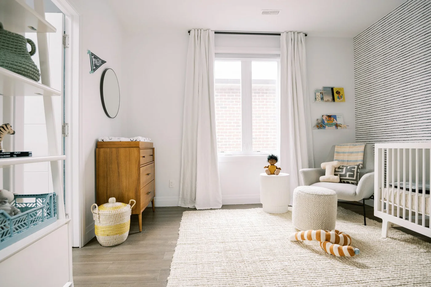 A pale nursery has fine dark horizontal wall lines beside a white crib, pale adult chair, wood drawer chest, round mirror and white curtains