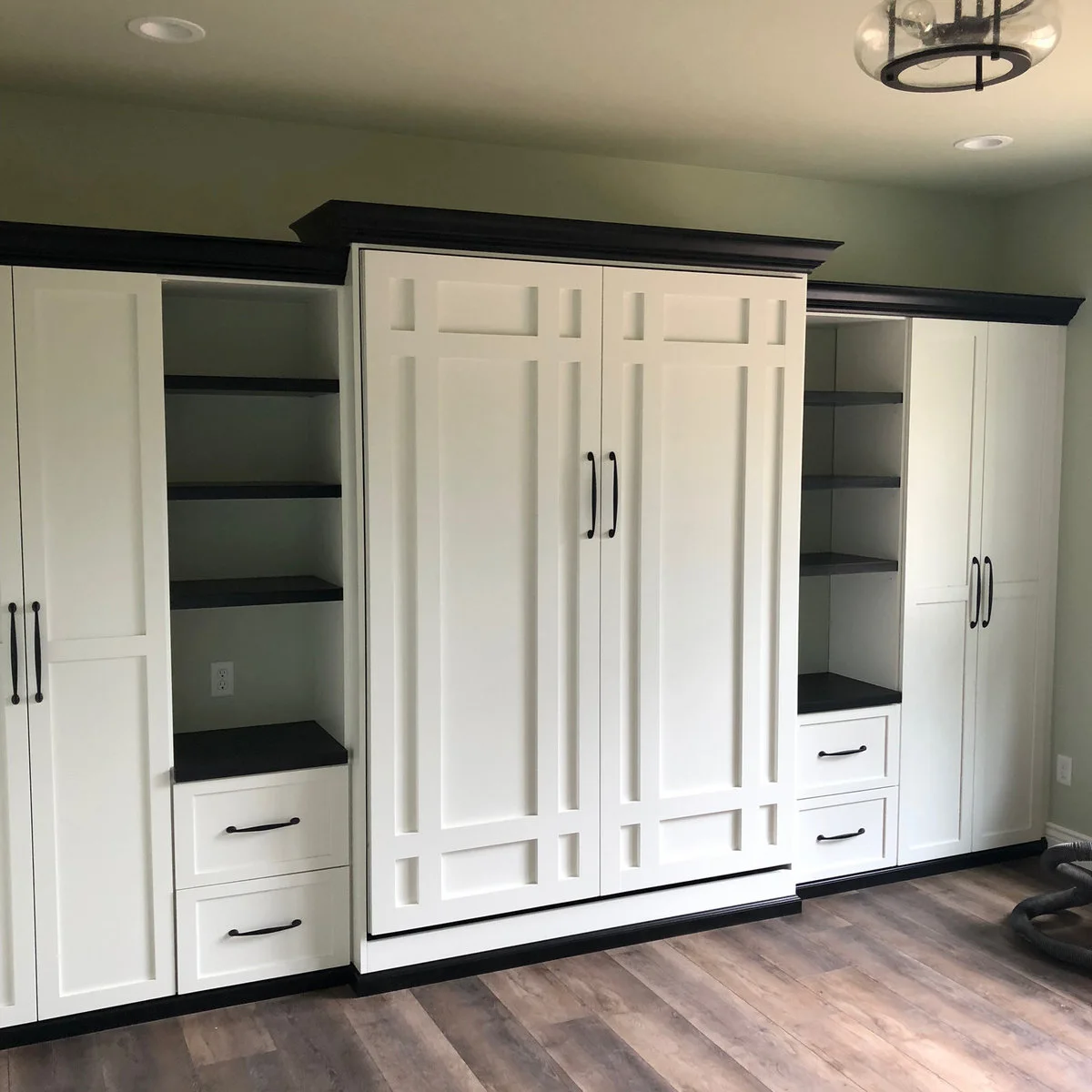 White built-in cupboards with dark trim and open side shelves surround a closed central wall-bed front in a shared sewing and guest room.