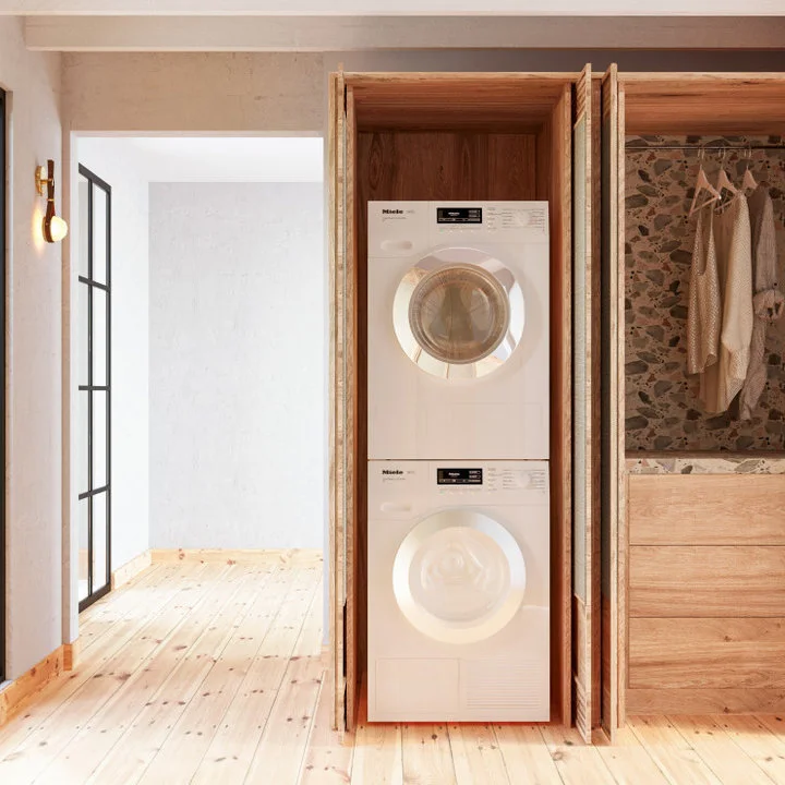 Pale wood folding doors open to stacked laundry machines beside a matching cabinet with drying rails.