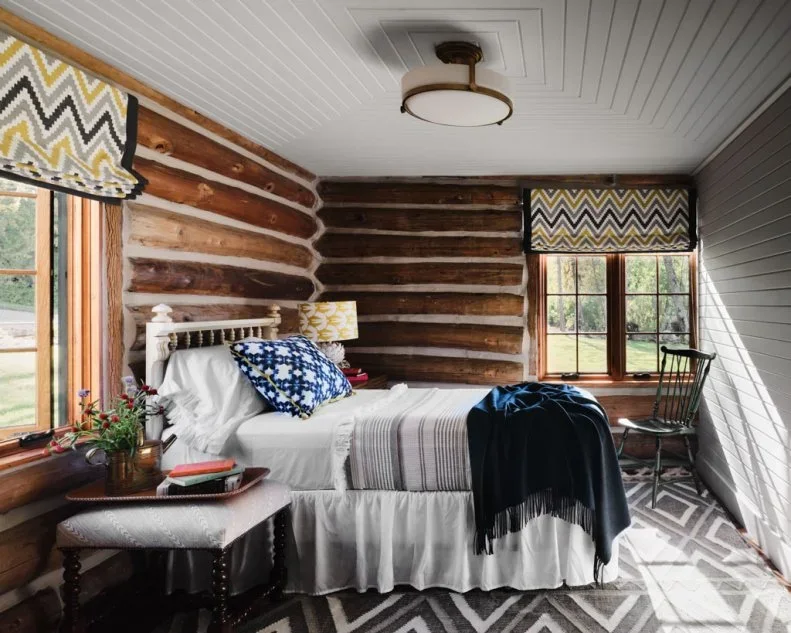 Dark log bedroom walls sit beneath a white boarded ceiling beside a narrow bed and zigzag window shades.