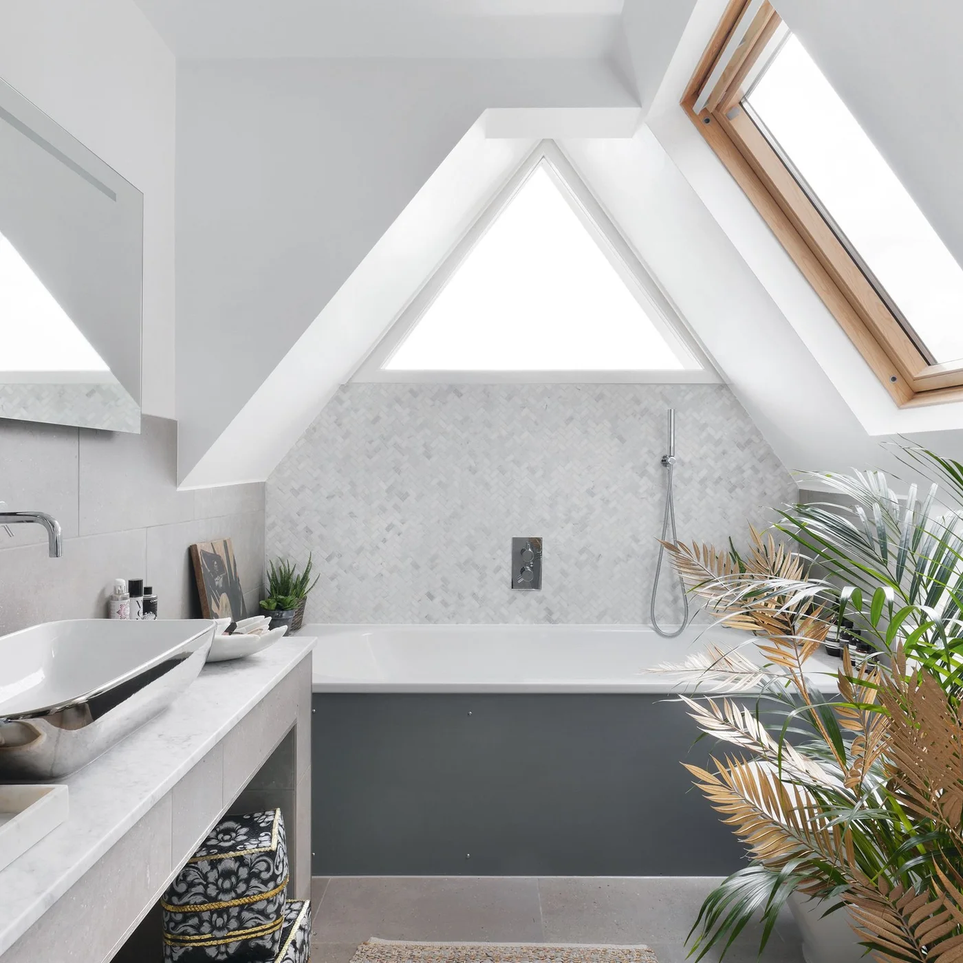 A compact loft bathroom places a gray-paneled bath beneath a sloped ceiling, beside a small wood vanity, triangular window and rooflight.
