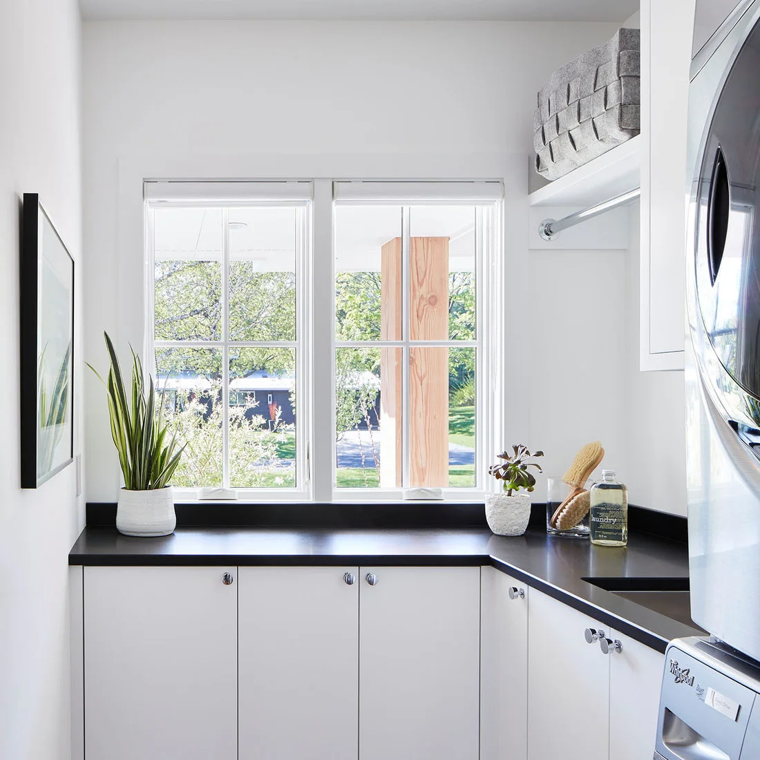 White flat-front cabinets and a dark L-shaped counter wrap below a window beside stacked machines and a high drying rail.