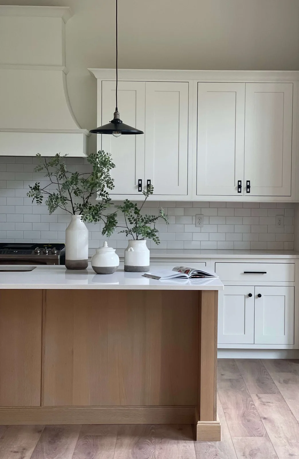 A white oak island with a pale countertop and paneled end stands before white cabinets with black pulls and a black pendant.