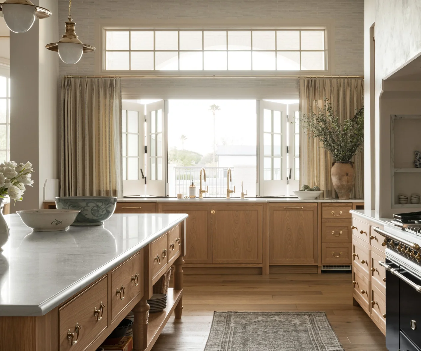 White oak lower cabinets and drawers run beneath an opening kitchen window, with pale counters, brass-colored taps and a patterned runner.