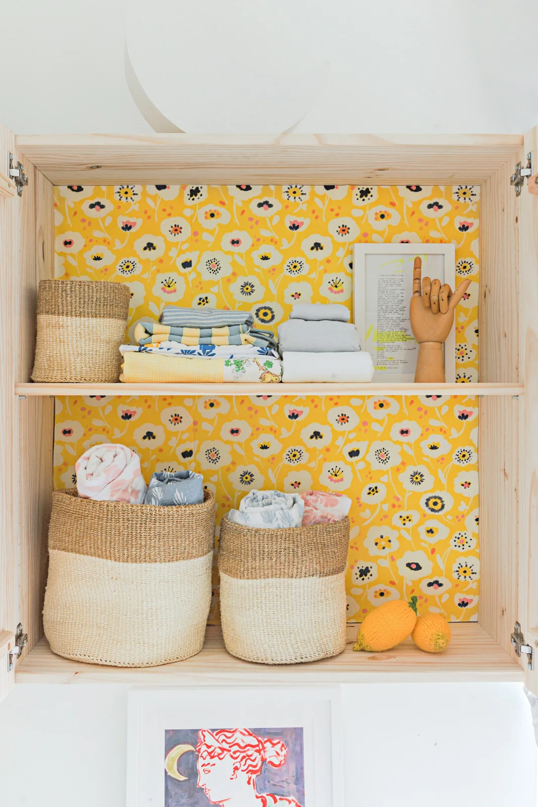 An open timber wall cabinet with yellow floral lining holds folded textiles and woven storage baskets.