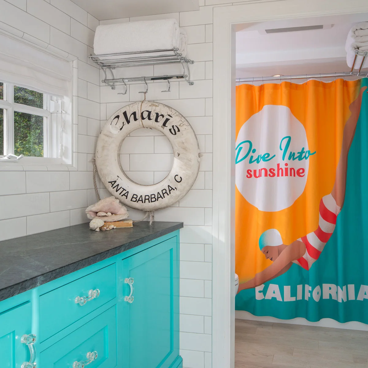 A bright turquoise cabinet with clear handles stands beside a colorful travel-style shower curtain and a wall-hung life ring.
