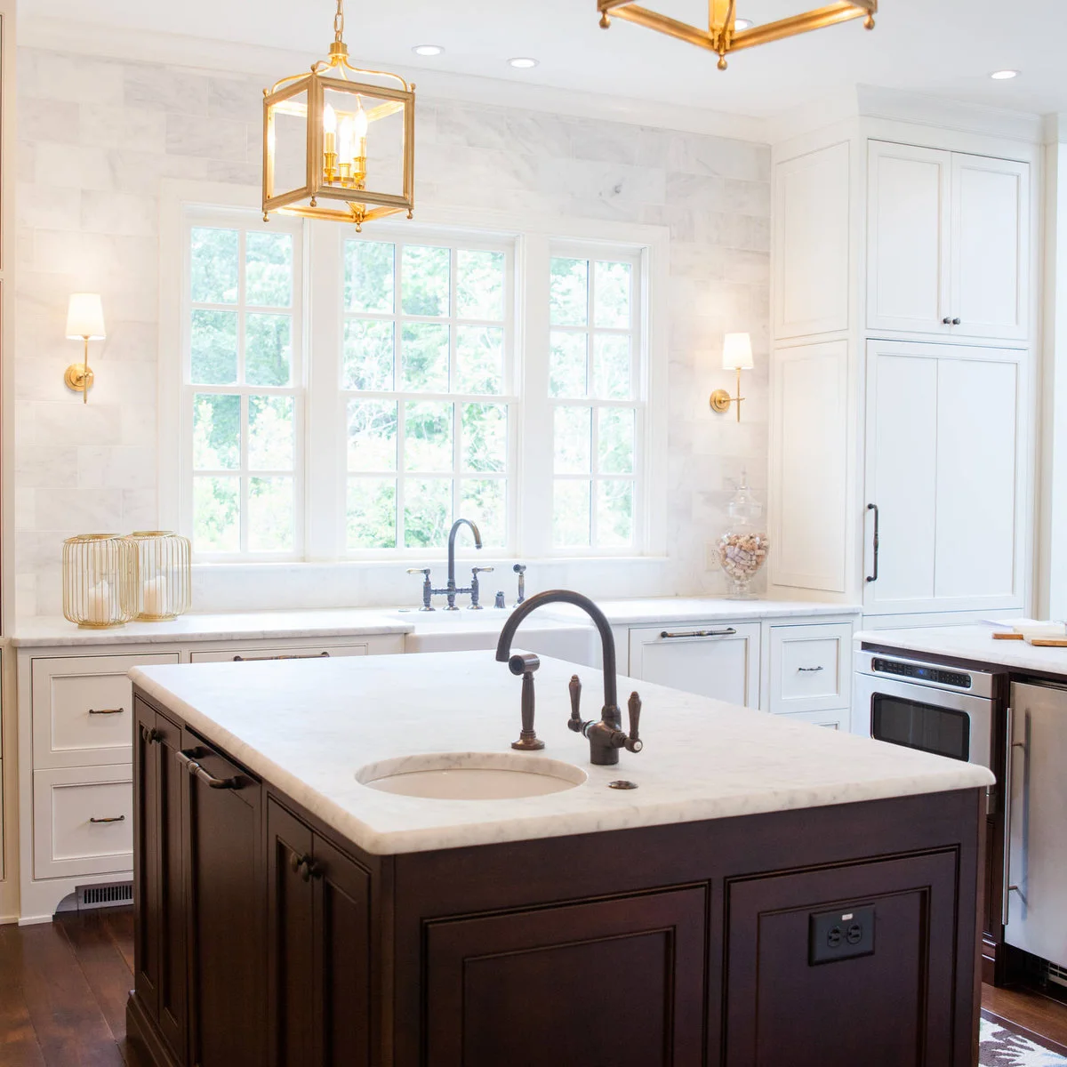 Two small pale shaded wall lights flank a wide kitchen window above a sink, with warm metal lanterns over a dark island.