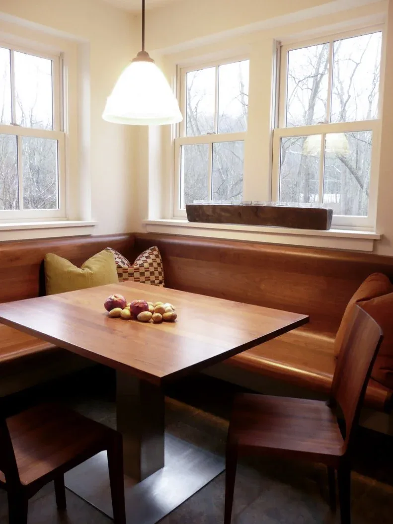 Wooden corner banquette beneath broad white-framed windows, with a square wooden table on a central metal support, dark wooden chairs, and two patterned pillows.
