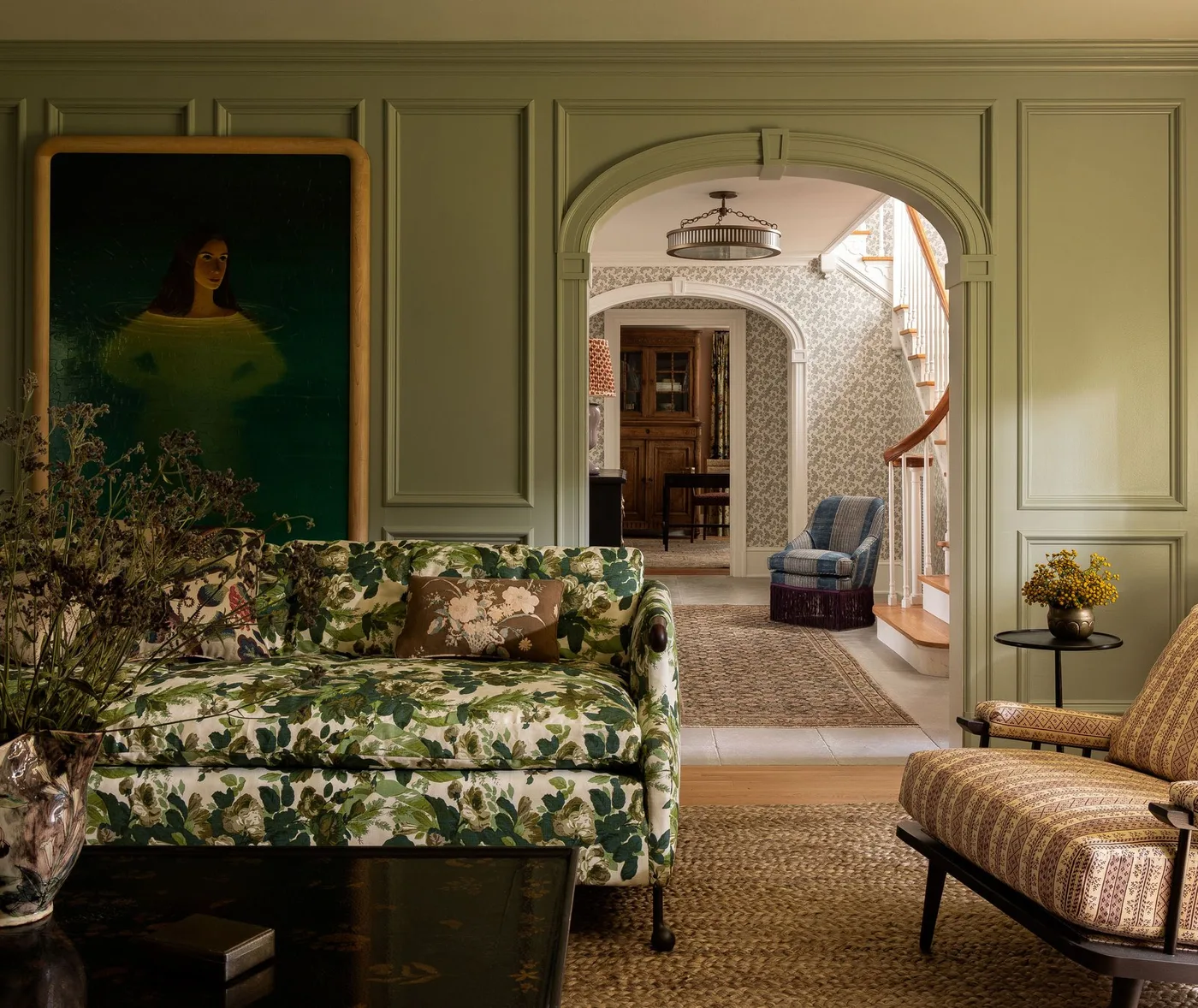 Muted green paneled living room with a botanical sofa, patterned armchair and dark round coffee table