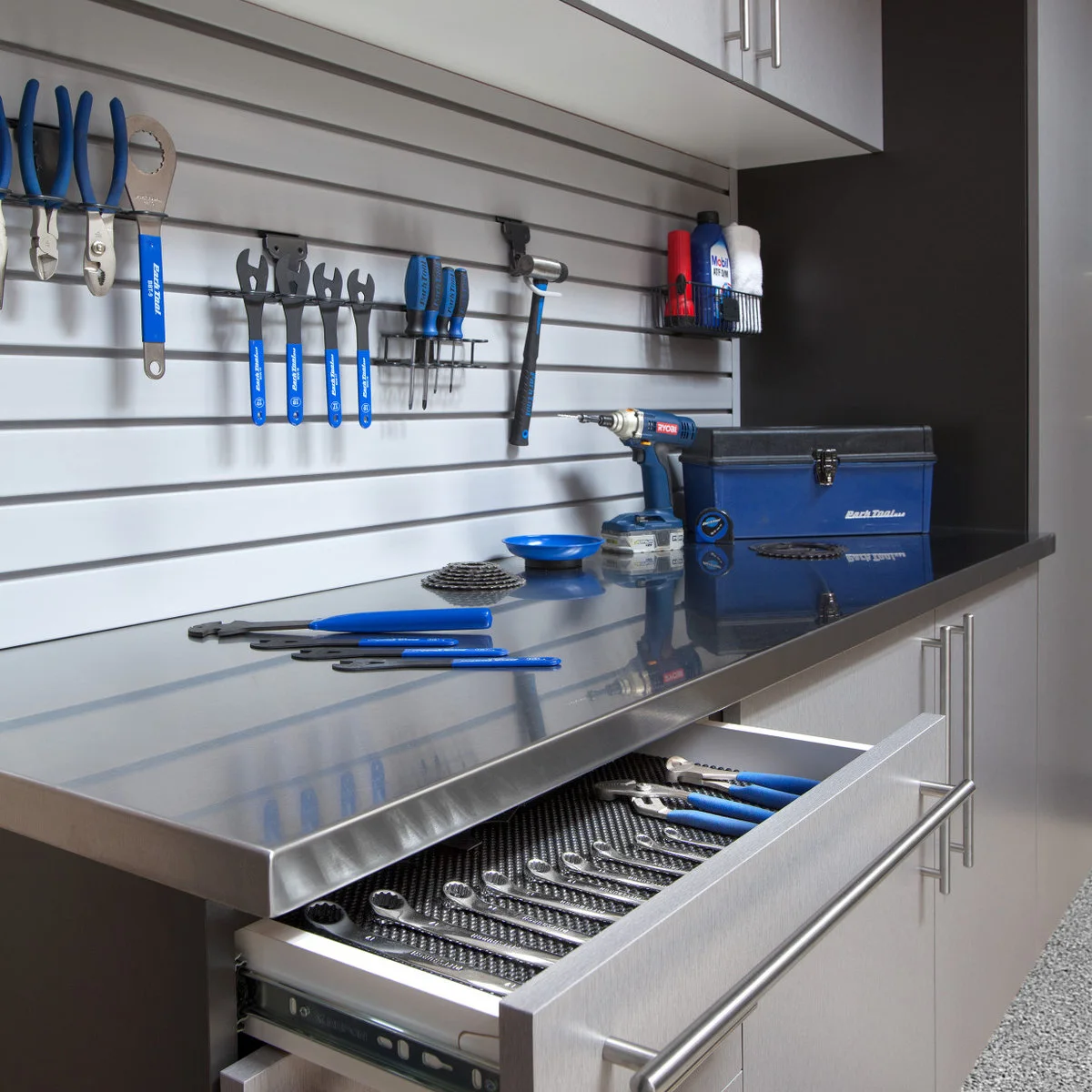 Garage workbench with a reflective metal top, an open shallow tool drawer and blue-handled tools on a slat wall.