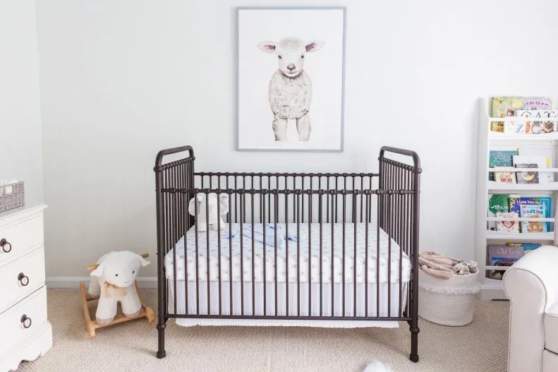Dark crib beneath lamb illustration in pale nursery with lighter storage, small rocking animal and adult seating.