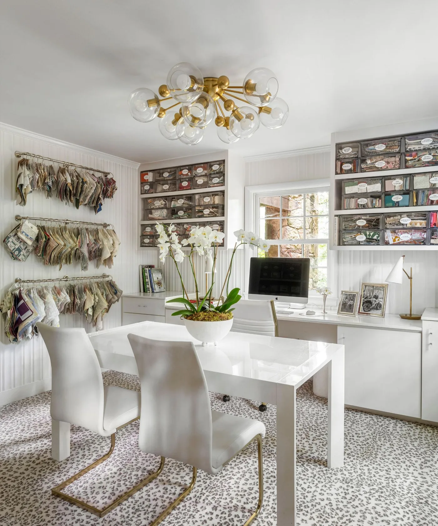 White office tables and chairs beneath a gold-toned globe ceiling light with shelves of boxes and hanging fabric samples.