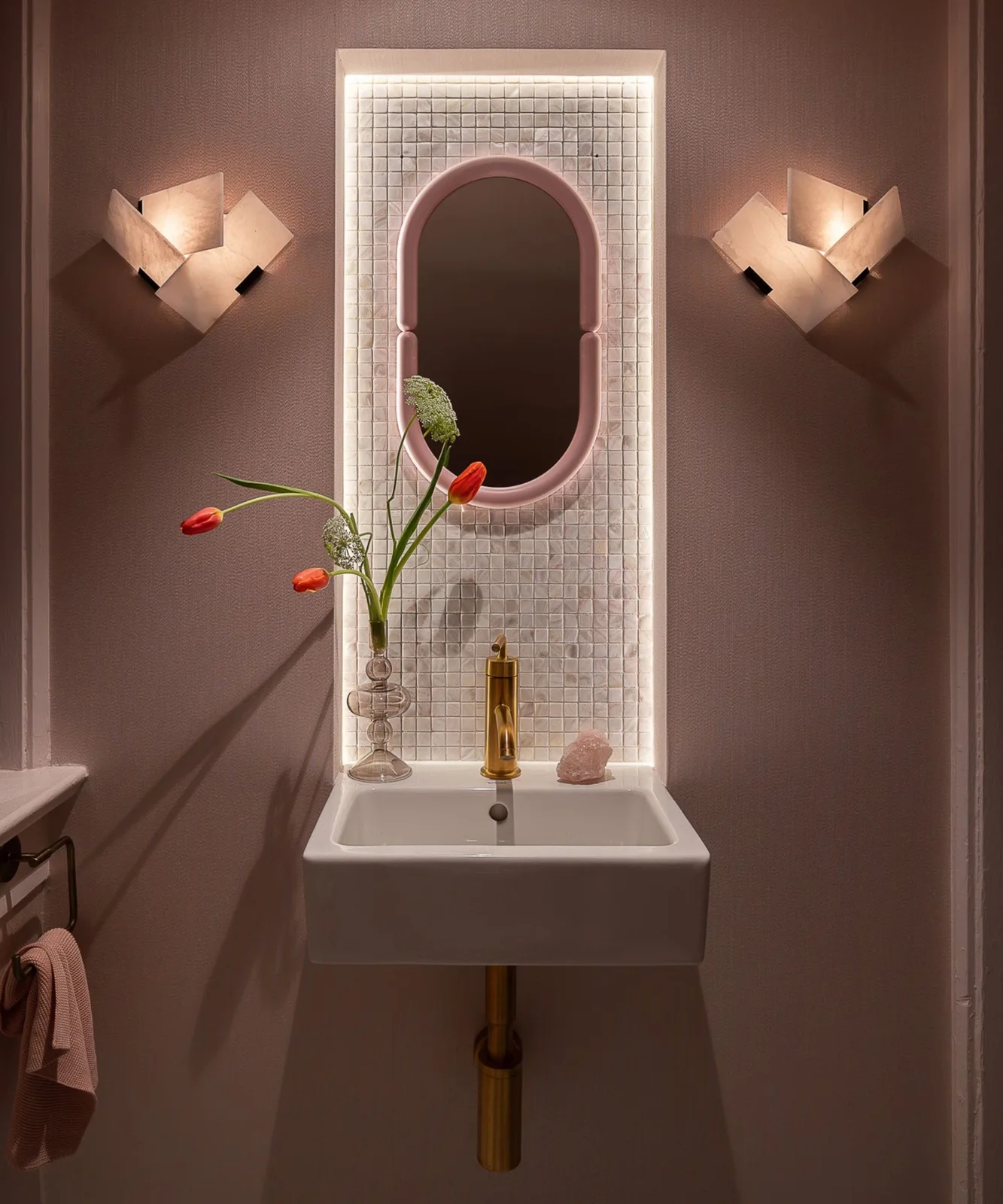 A compact white wall basin stands below a pink rounded mirror in a lit pale tiled recess between sculptural wall lights and plain pink walls